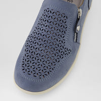 A close-up of a slip-on shoe in dusty blue suede with geometric laser-cut perforations on the toe box, a side zipper with silver metal hardware, and a cream-colored sole.