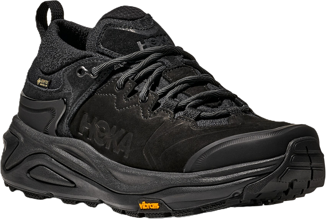 A black Hoka hiking shoe with a thick black sole, featuring the Hoka logo on the side and a small orange Vibram logo on the outsole.