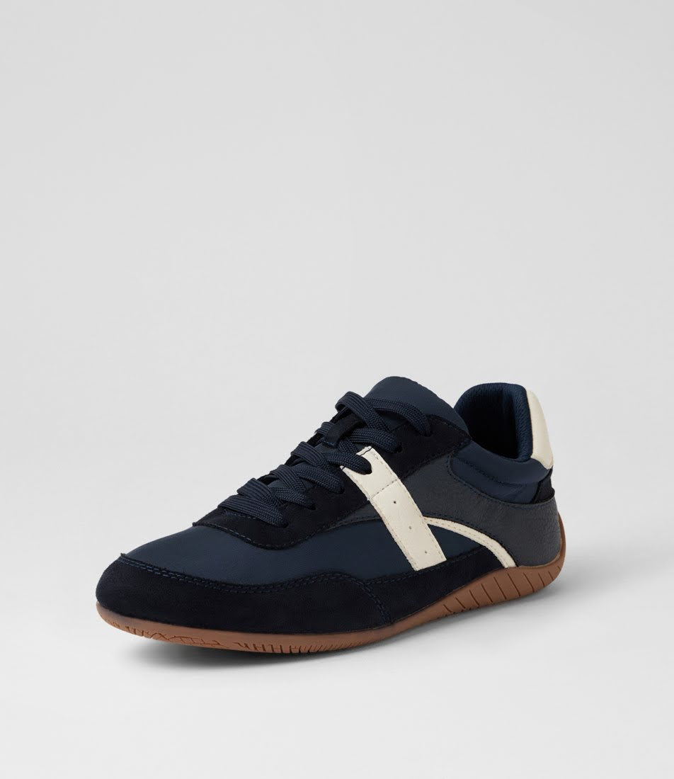 A low-top sneaker in navy blue suede and synthetic fabric, featuring a contrasting off-white side stripe, navy laces, and a brown gum sole, shown against a light gray studio background.